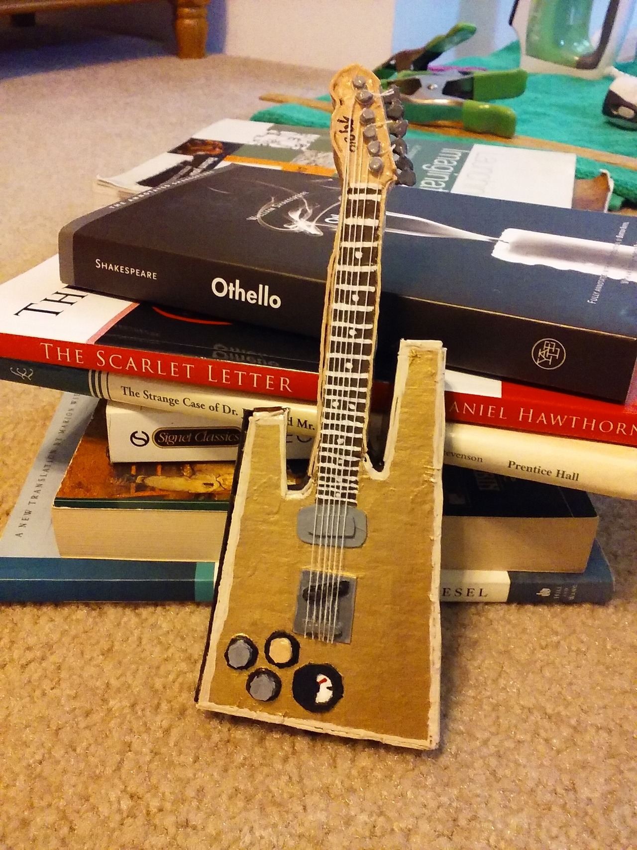 The final cardboard miniature Chessmaster. It is a golden guitar with a wooden neck. There are some knobs in the corner of the body and also has some sort of dial. It is leaning against a stack of books.