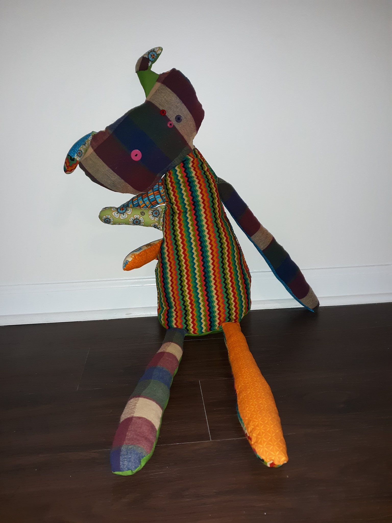 A very large doll sitting on the floor. She has a bean shaped head that is of a plaid pattern. She has two ears sticking out the top that are boomerang shaped and are much smaller than her head. She has one pink button on the left side of her head and three buttons on the right side of her head that are red, pink, and purple. Her body is very long and gets slightly wider as you reach the bottom. The front of her body is of a colorful chevron pattern. She has three short arms on the left side of her and one long arm on the right side of her. The three short arms each have a different pattern on them with the top one having a square pattern of browns, blues, and oranges, the middle arm having a green fabric with blue flowers, and the bottom arm being orange. The arm on the right side is of the same plaid fabric as her head. She has two long legs. The leg on the left is plaid and the leg on the right is orange.