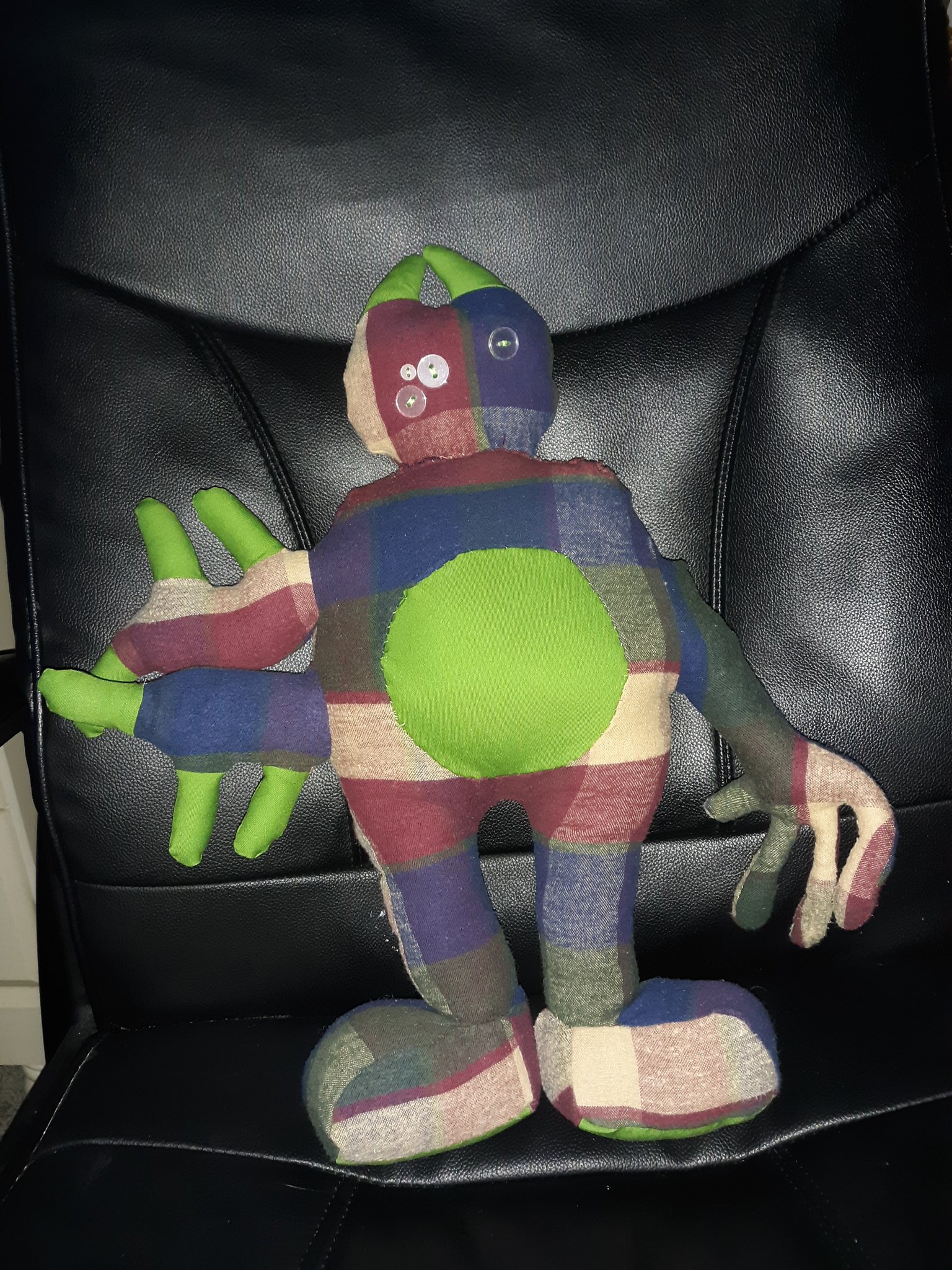 A green and plaid doll sitting on a black chair. The doll has a small plaid round head with two green horns coming out of it. It has four eyes made of clear buttons. The right side of their head only has one button and the left side has three buttons. The doll has a large body made of the same plaid material. It has a round green belly. They have two arms sticking out of the left side of their body. Each arm has three green fingers like eyes on a potato. The right side of their body only has one arm that has four fingers on it. They have two legs with rectangular feet.