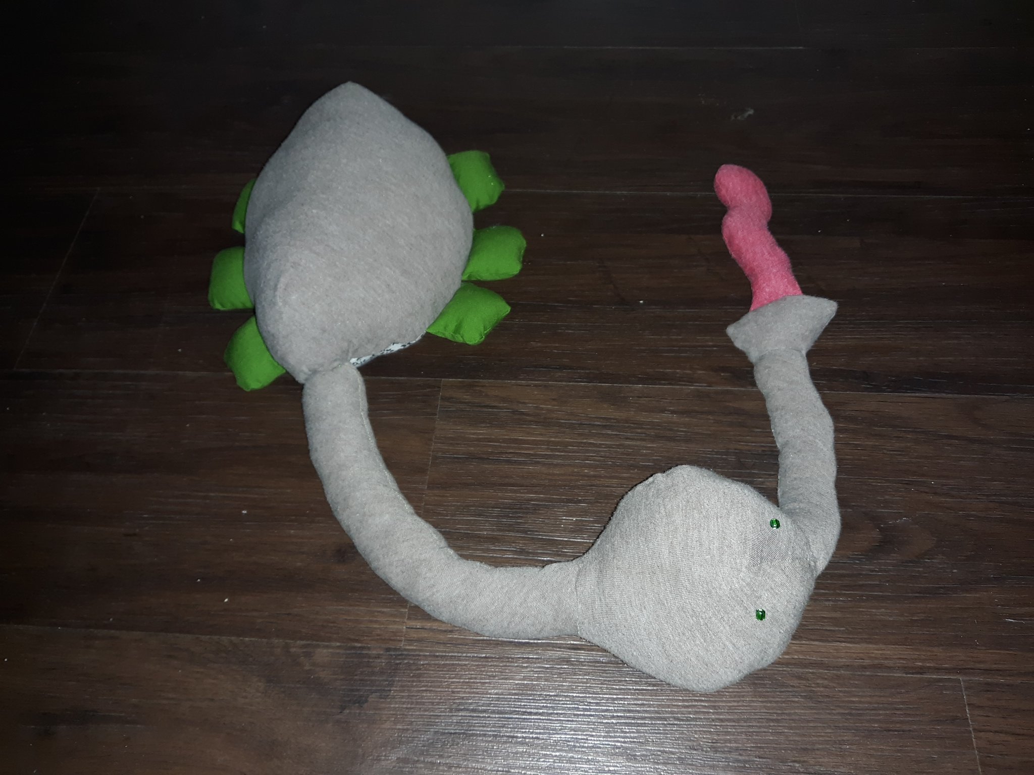 The top of a grey doll laying on the floor. It has a round grey body with a very long neck with a round head at the end of it. The head has a long snout coming out of it with a long red tongue coming out of that. The head has two small green beads for eyes. The body has six stubby green legs with three on each side.