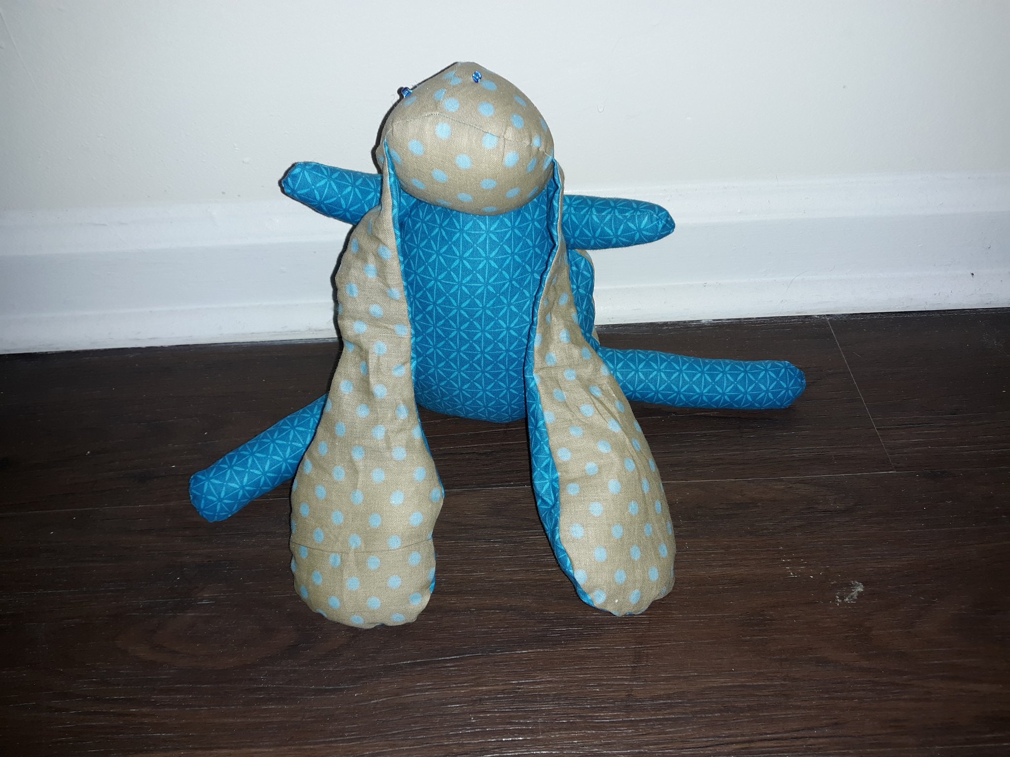 A blue and beige doll sitting on the ground. They have a small round head with long, floppy ears that touch the ground. Their body is larger than their head, but shorter than their ears. They have four short legs coming out of their body. Their head is of a beige fabric with light blue polka dots. They have two tiny blue beads for eyes. The top of their is also made of the same fabric, but the underside is of a blue fabric that has a lighter blue pattern on top. Their belly and legs are made of the same blue fabric.