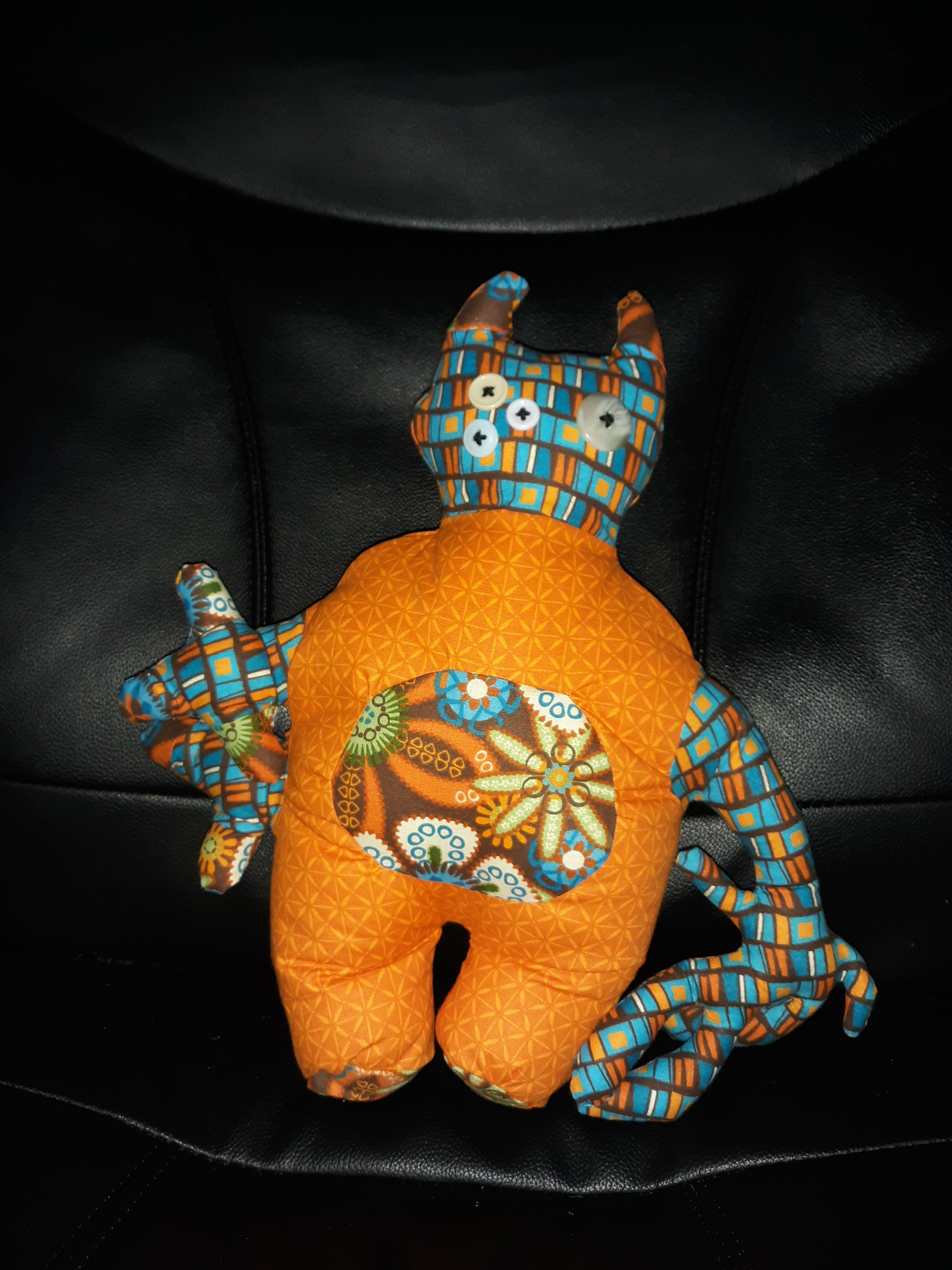 An orange doll standing on a black chair. They have a small head consisting of a brown, blue, yellow, and orange rectangle pattern. They have two horns sticking out and have three button eyes on the left of their face and one large button eye on the right of their face. Their body is orange with a brown floral pattern belly. They have two short stubby legs that are also orange. They have two short arms on the left side of their body and one very long arm on the left side of their body. Both arms have that rectangle pattern on them.