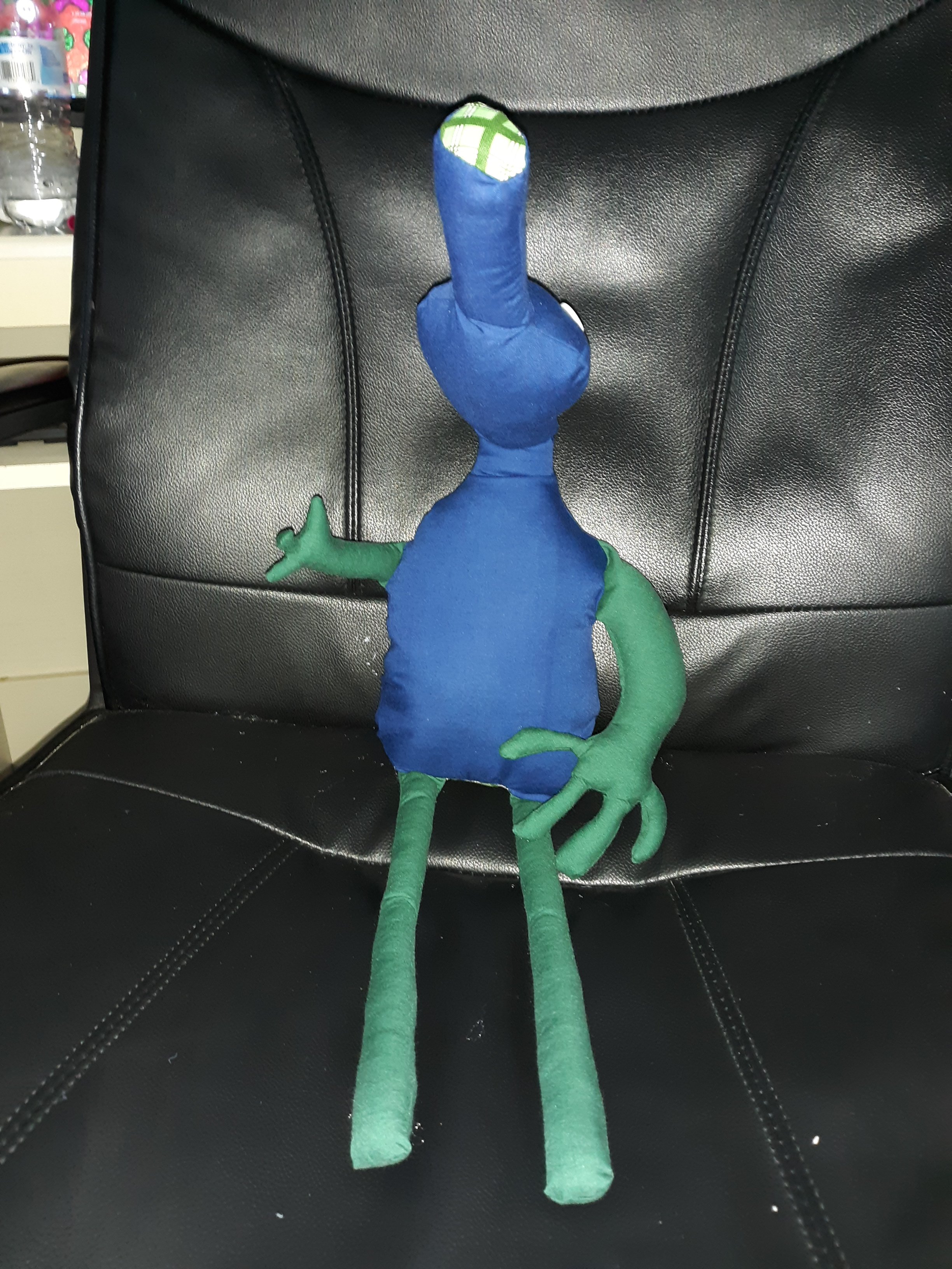 A blue and green doll sitting on a black chair. They have a small head with a long snout that has a plaid green pattern at the end of it. The head has one large button for an eye on the left and two smaller button eyes on the right. The head and the body are all dark blue with the arms and legs being dark green. They have two arms with the left arm being very short and small and the right arm being long and large. The two legs that they have are thin and long.