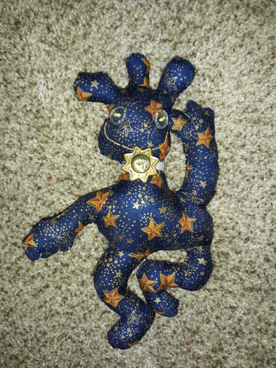 A doll with a celestial print on it. The background is dark blue with golden stars all over. It has a head with three short growths coming out of it. They have two glittery, clear button eyes with a yellow embroidered smile. They have a white ribbun with a sun button wrapped around their neck. They have two arms with one facing down and one stretched upwards as if they are waving. They also have two bent legs that look like they're dancing.