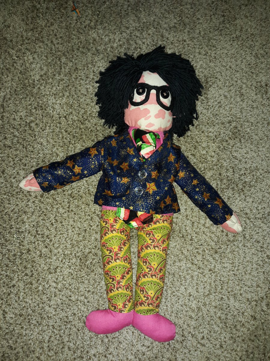 A doll of a fictional character named Leslie Down. He is a white man with red scars all over his body. He has black yarn for hair and is wearing black glasses over his black button eyes. He is wearing a pink button up with a celestial blazer on top. He is wearing a tie that has a pattern of sweets on it. His pants are of an intricate pattern consisting of green, yellow, brown, and red. His shoes are pink.