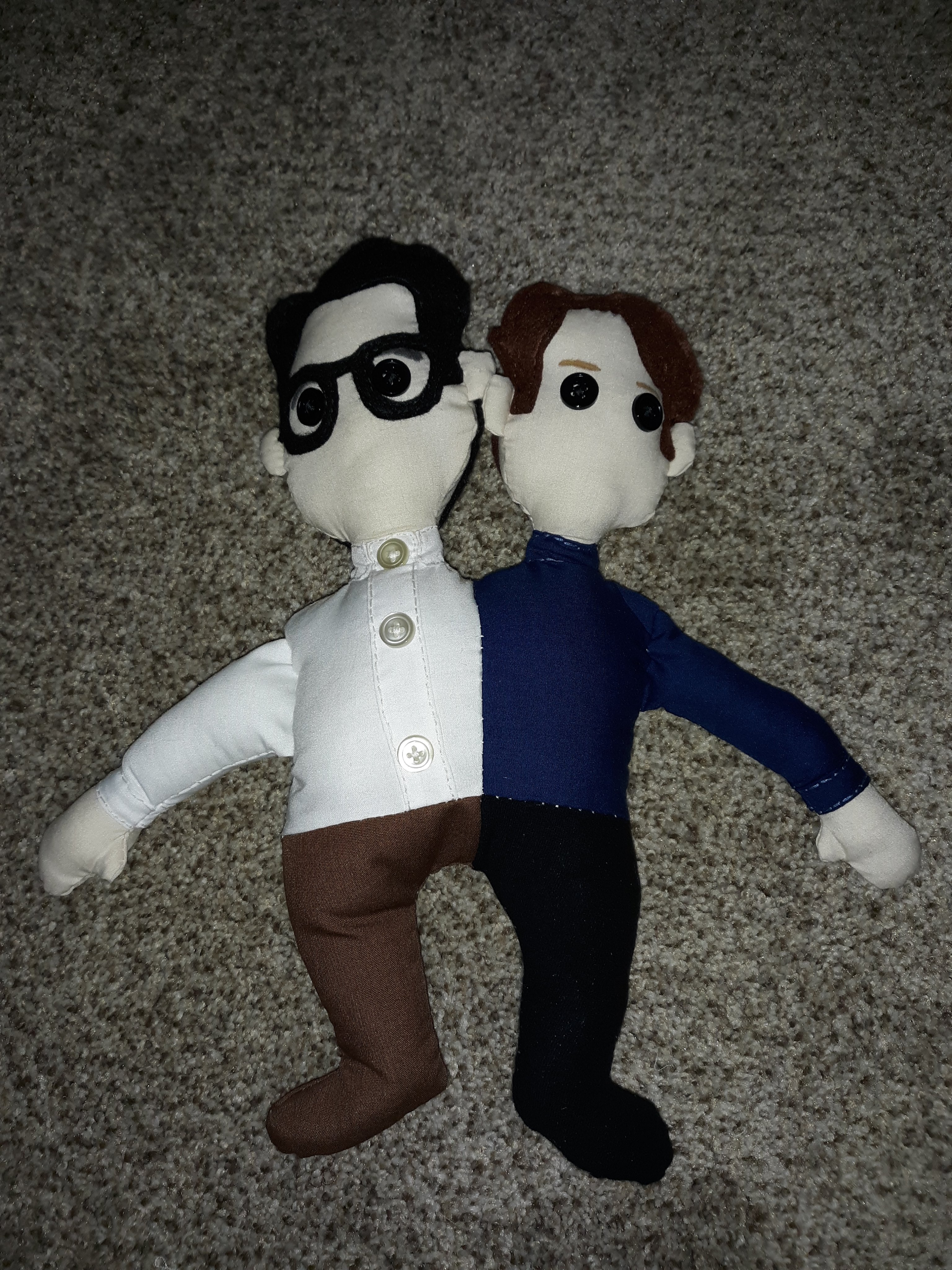 A doll of a young John Flansburgh and a young John Linnell sewn together as one. Flansburgh is on the left side wearing a white button up and brown pants. He has three buttons going down his shirt. He has short black hair and is wearing black glasses. He has two black buttons for eyes. Linnell is on the right side and is wearing a dark blue turtleneck with black pants. He has short brown hair and has two black buttons for eyes.