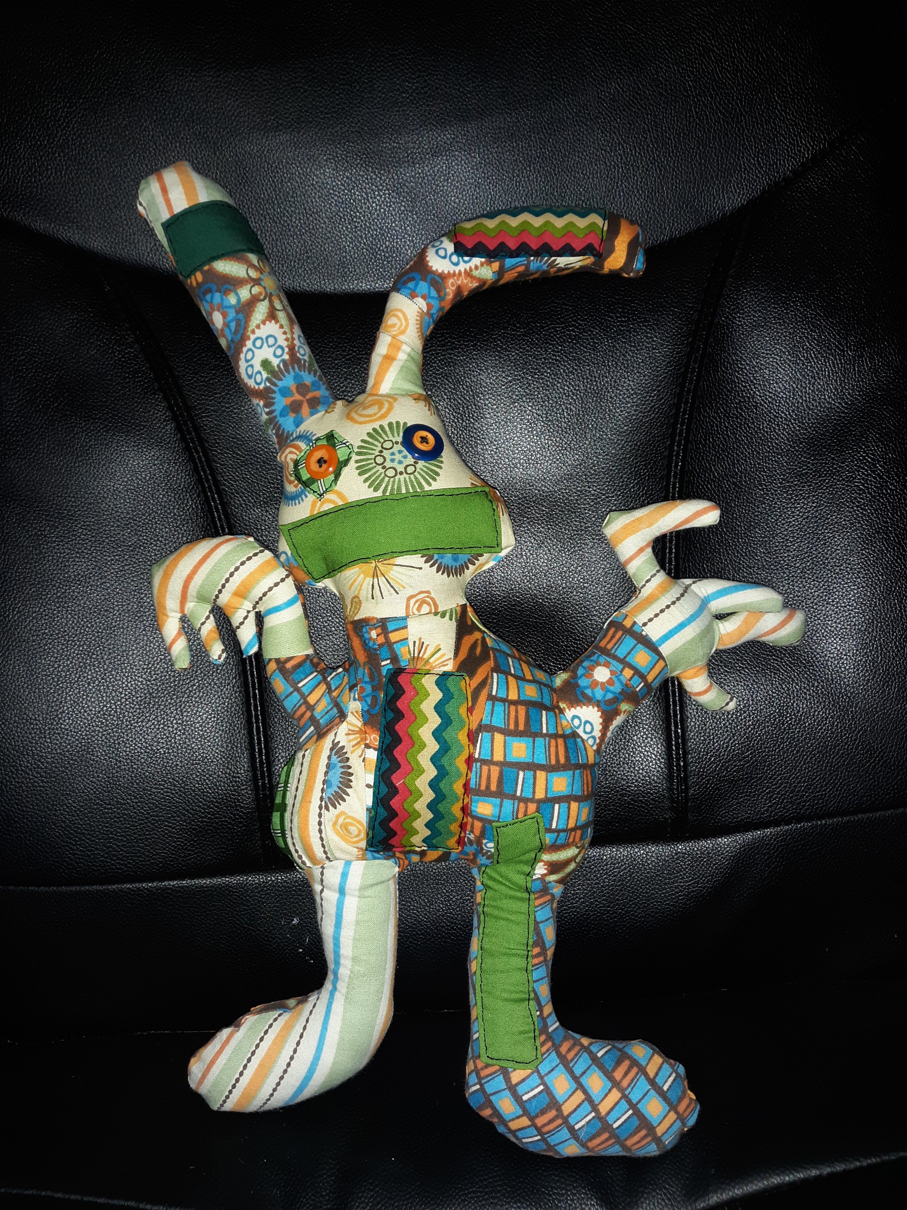 A patchwork doll that looks somewhat like a rabbit. It has two long ears sticking out of a head that has two orange and blue button eyes. There is a green mouth sewn onto the face. The body is somewhat triangular and has two arms sticking out with odd shaped hands. The doll also has two legs with rounded feet. The doll consists of different patterns like chevron, tiger, stripes, abstract floral, solid greens, and some rectangles.