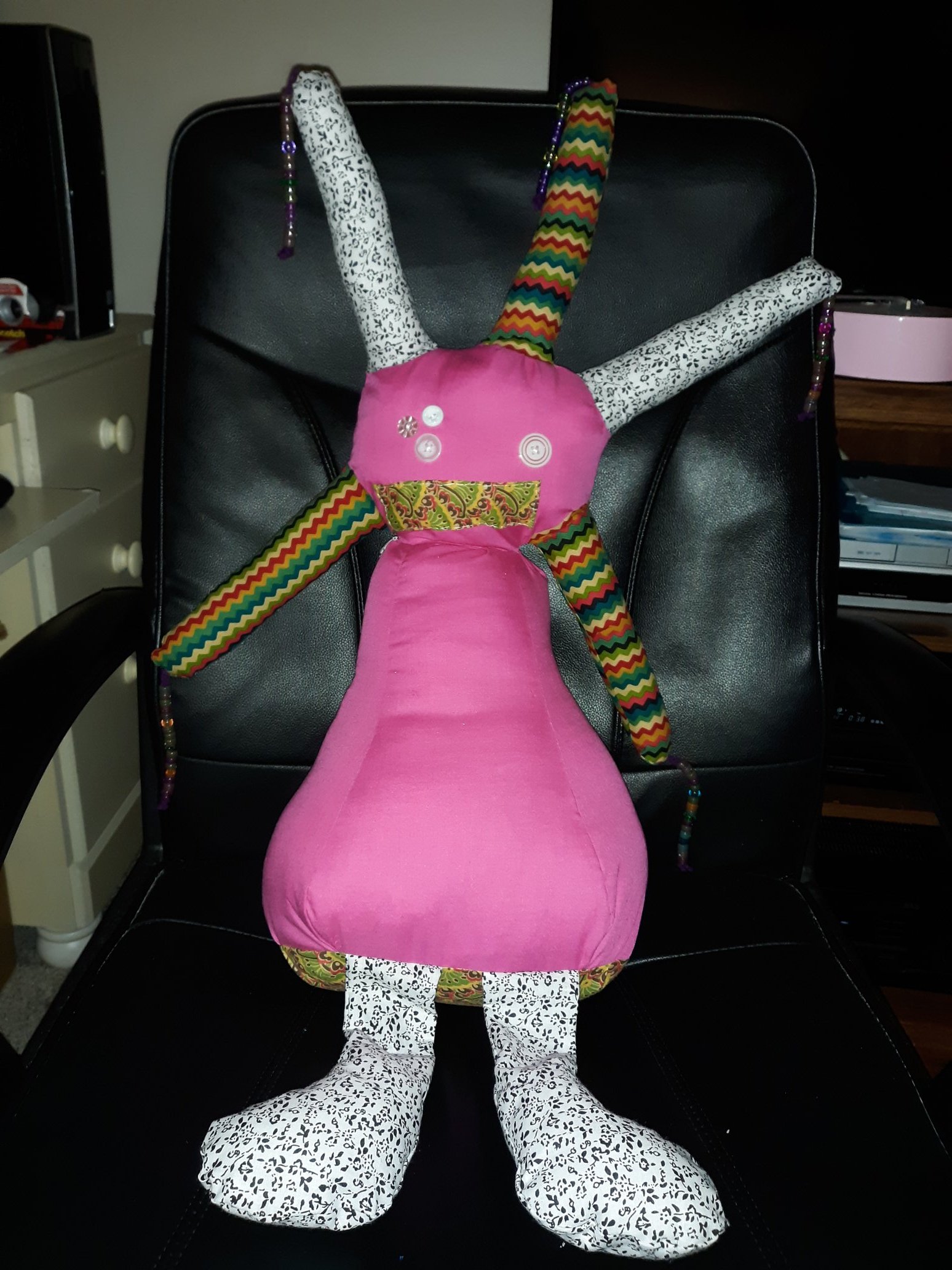 A very large pink doll. It has a circular head with five growths coming out of it. Each growth has a string of beads hanging off the ends of them. Three of the growths have a colorful chevron pattern and two of them have a black and white floral pattern. The face of the doll has three white button eyes on the left side and one white button eye on the right side. The mouth is a rectangle piece of fabric sewn on. The body is much larger than the head. It gets larger as it goes to the bottom. The bottom of the doll is of an intricate pattern and there are two legs sticking out that are of the same black and white floral pattern.