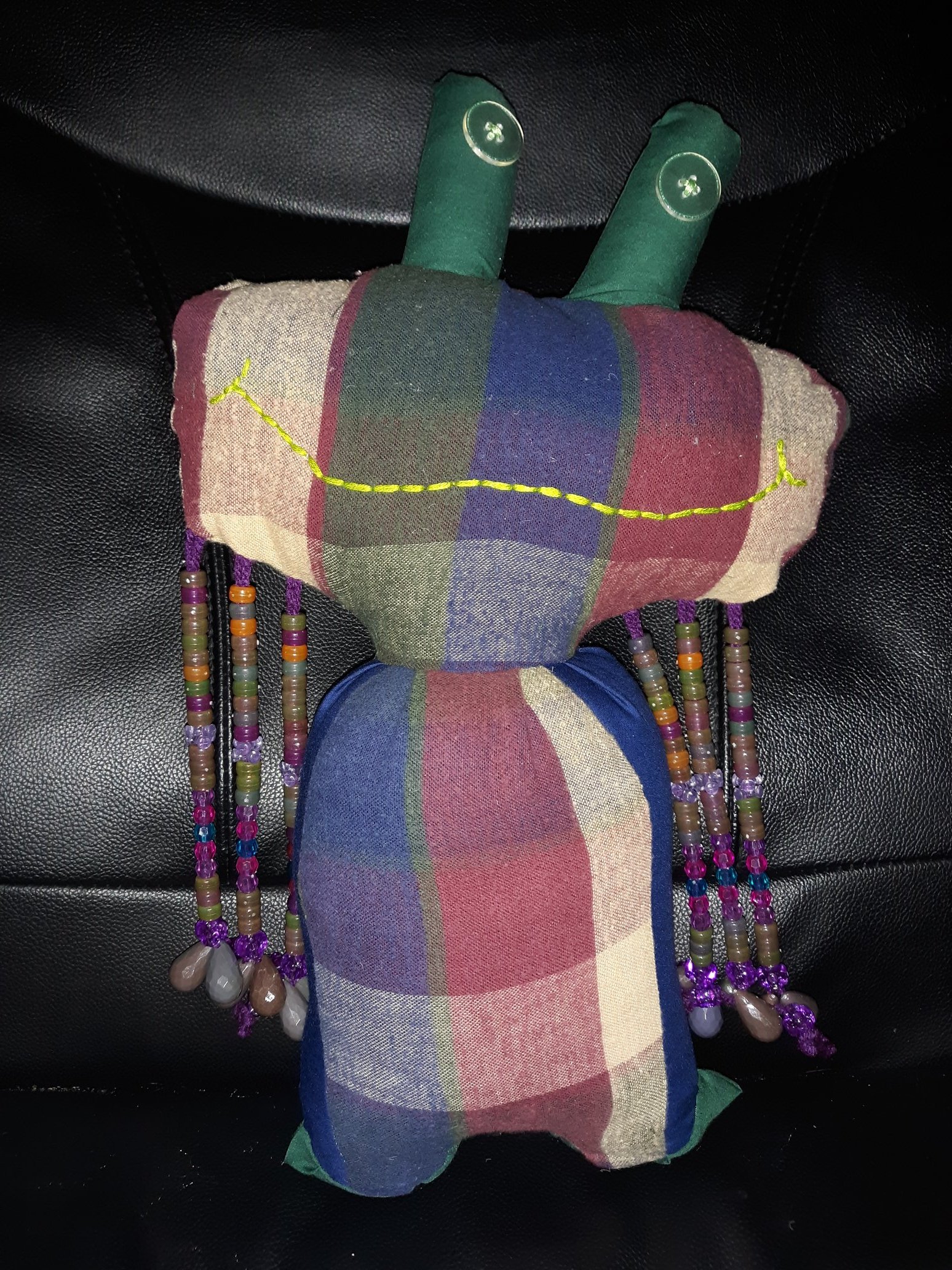 A doll that has a large, wide, plaid head with two little green cylindrical shaped growths coming out of the top. Each cylindrical shaped growth has a clear button eye sewn onto it. The head has a wide, highlighter green smile. There are three rows of beads hanging down from each side of the head. The body is also plaid with dark blue sides and the same green as the cylindrical growths on the bottom where the stubby feet are.