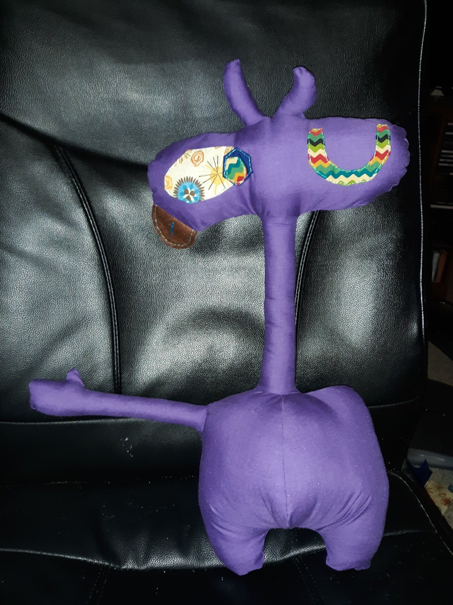A purple doll with a pill shaped head and a very long thin neck and a round body that has one thin arm sticking out it. The head has two small purple horns coming out of the middle of it. The face is of two eyes with one winking. It has a brown tongue sticking out of the left side of the face. The arm has one hand with the thumb sticking up as if they are asking for a high five from you. They have two stubby feet at the bottom of their body.