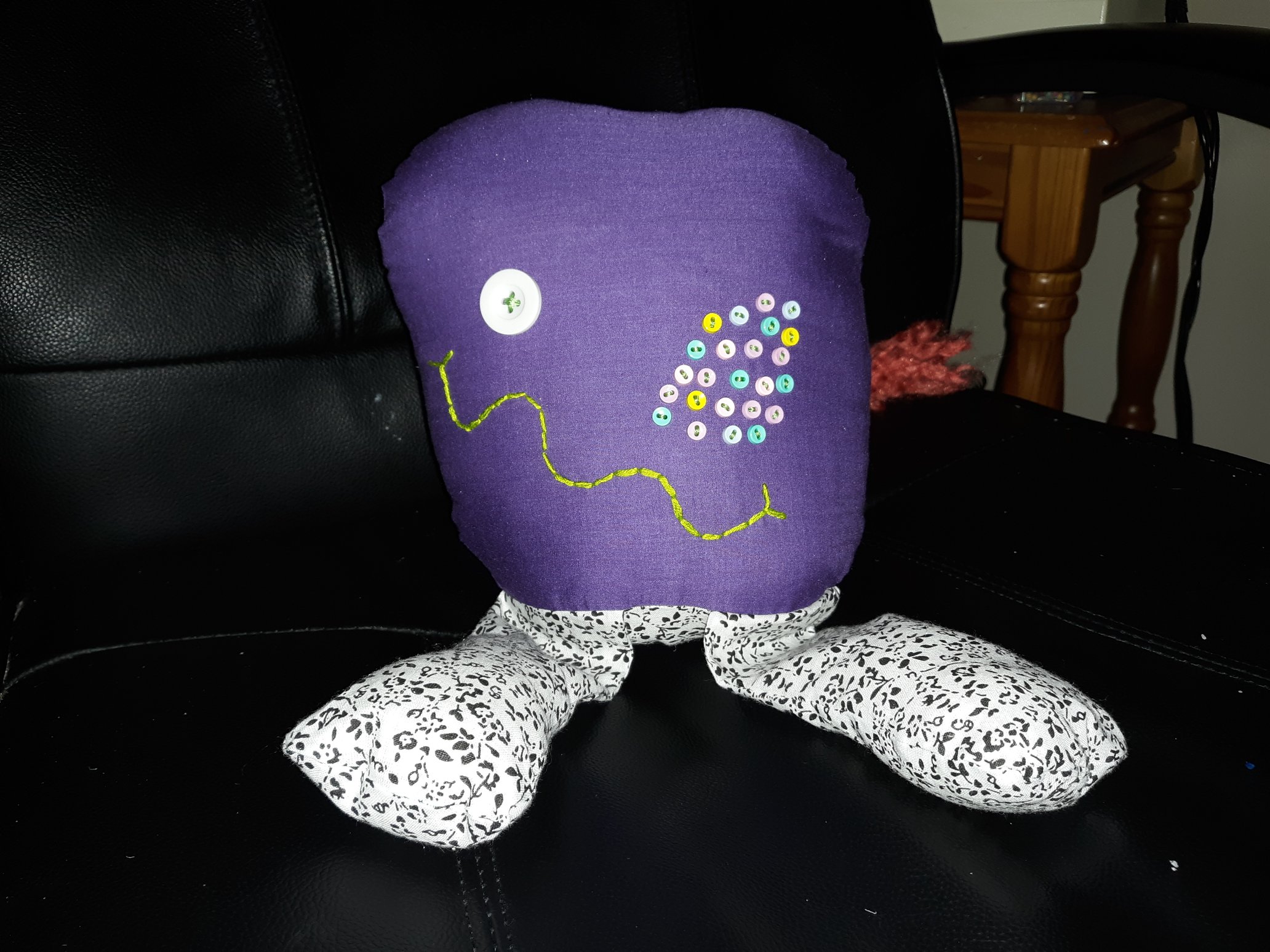 A very round purple doll with many eyes. It has one white button eye on the left of the face and many smaller button eyes on the right side of the face. The smaller button eyes are purple, pink, yellow, and blue. It has a wide squiggly green smile on its face. There are two legs coming out the bottom of it that are of a black and white floral pattern.