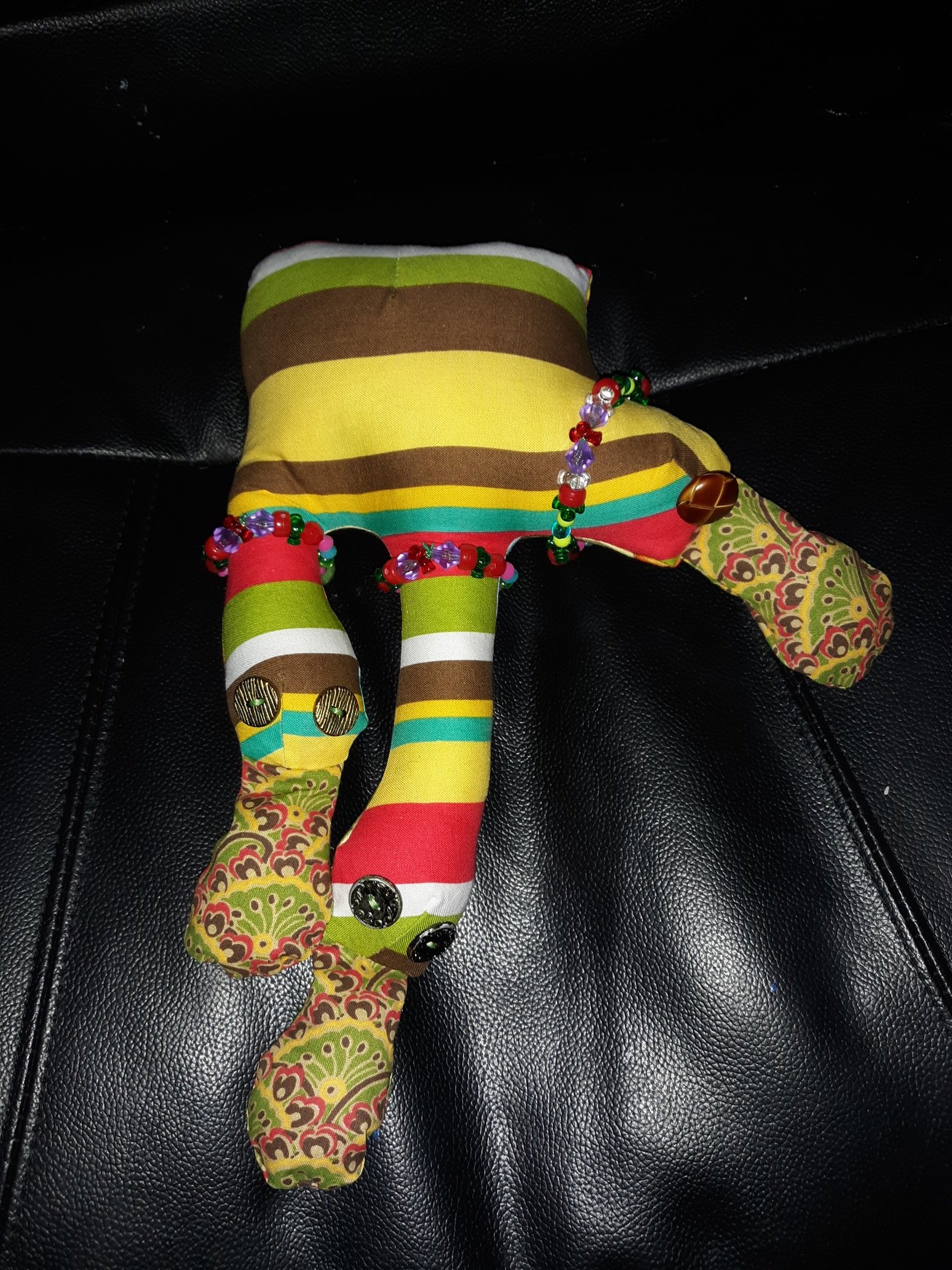 A three headed doll. The body is mostly square shaped with three different sized heads coming out of it. The head on the far left has a thin neck with a larger round head coming out. It has two button eyes and a large tongue sticking out. The head in the middle has a longer neck with a round head coming out of it. It also has two button eyes and a tongue sticking out. The last head on the right is much shorter with only one button eye and a tongue sticking out. The body is a striped fabric with their tongue having a more intricate pattern on it. Each head is wearing a beaded necklace.