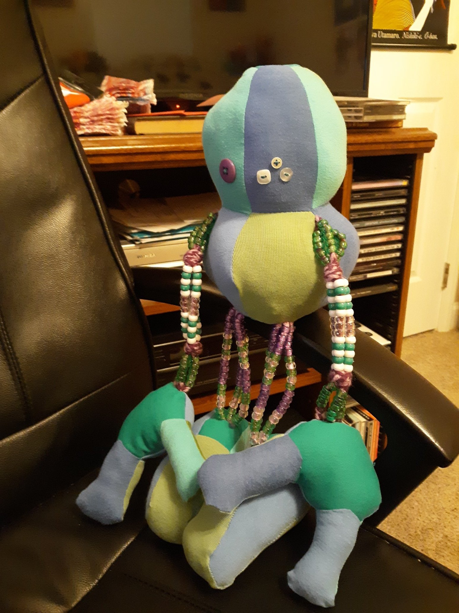 A doll sitting on top of the arm of a chair. The doll is made of different shades of blues and greens. It has a round head with a round body. The head has one purple button eye with three smaller white button eyes. They have two arms sticking out of the sides of their body. The arms are made of white and green beads. The ends of the arms have hands that are somewhat like claws. The doll also has two legs coming out of the body at the bottom. The legs are also made of beads like the arms and each have a foot at the ends of them. The feet are shaped somewhat like a trapezoid.