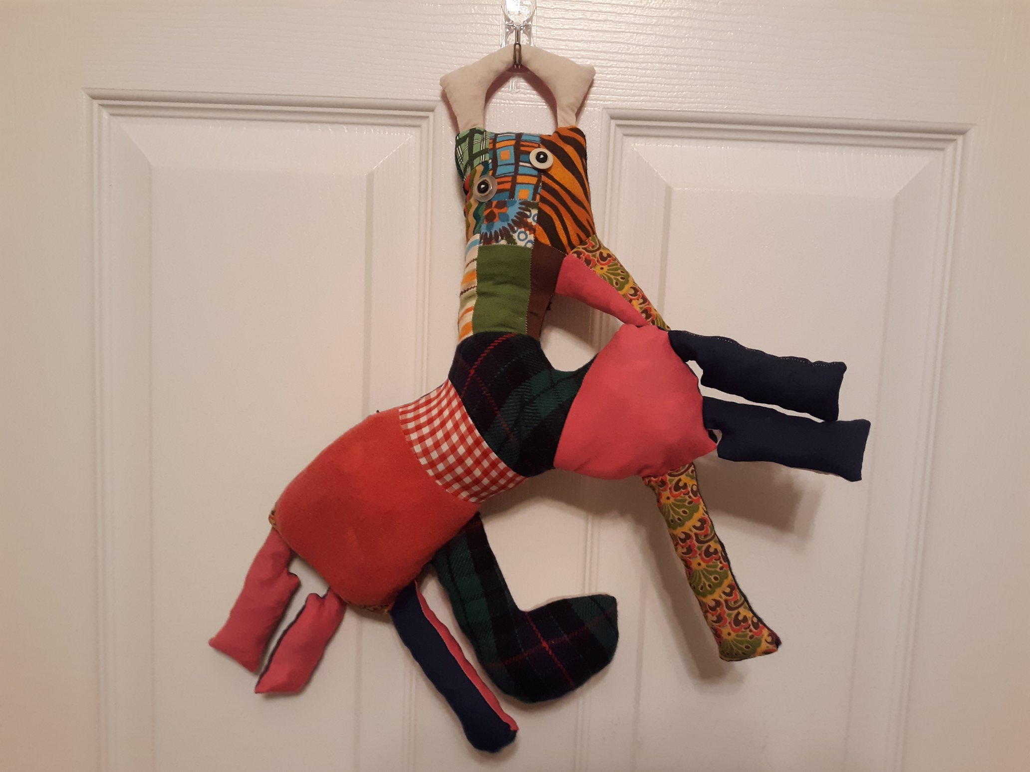 A patchwork doll hanging off of a hook that is attached to a door. The doll has a rectangular head with a triangular beak on the side of the head. There is a handle on the top of the head. The doll has two button eyes. The head comes out of the center of the wide body of the doll. There are two legs popping out horizontally of the butt of the doll with two more legs vertically coming out of the bottom. At the front of the doll's body are three more legs. One leg is coming out vertically from the bottom and two legs are coming out horizontally from the side.