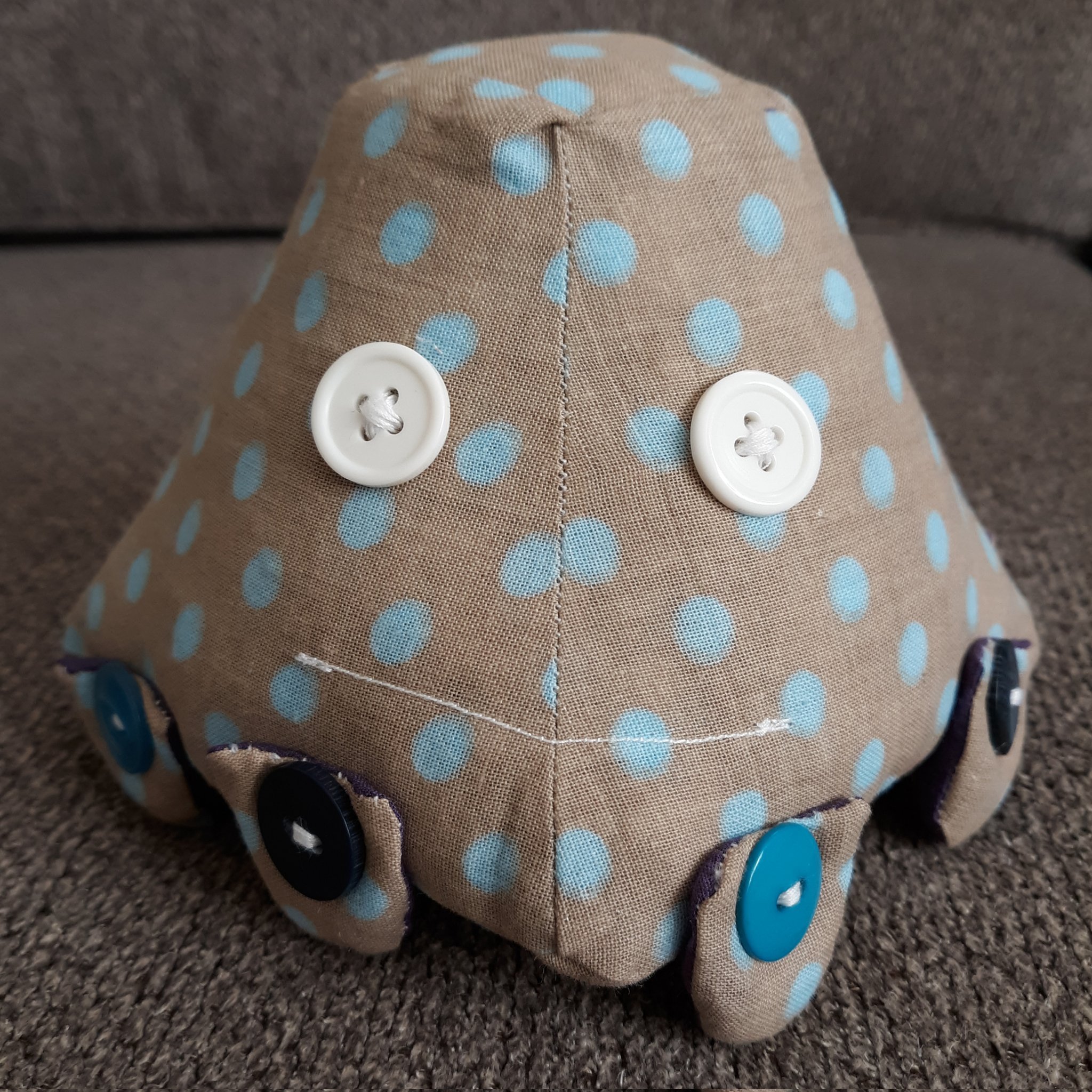A small pyramid shaped doll. It has four different sides with a square top and square bottom. The body is of a brown fabric with blue dots on it. The eyes are made of two white buttons with a white mouth sewn onto it. The doll has eight legs, two on each face of the body, that are sewn on with blue buttons.