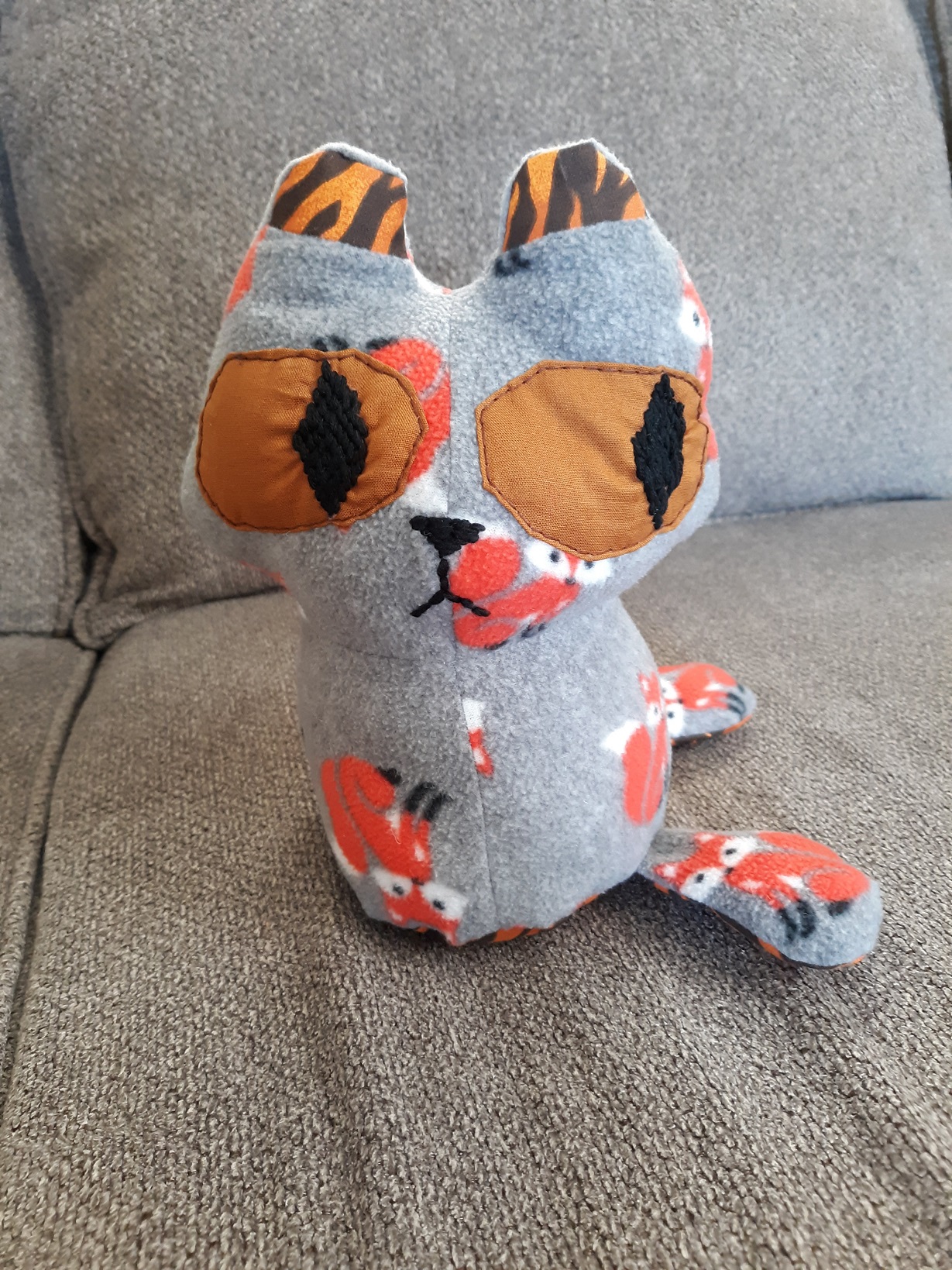 A doll that looks somewhat of a cat. Their body is made of a grey fabric that has orange foxes on it. The inside of the ears are of a tiger print. The eyes are large and brown with black diamond shaped pupils that were embroidered on. The mouth and nose were also embroidered on. The doll has two legs that are sewn on the side of the body. The bottom of the body and the bottom of the legs have a tiger pattern on them.