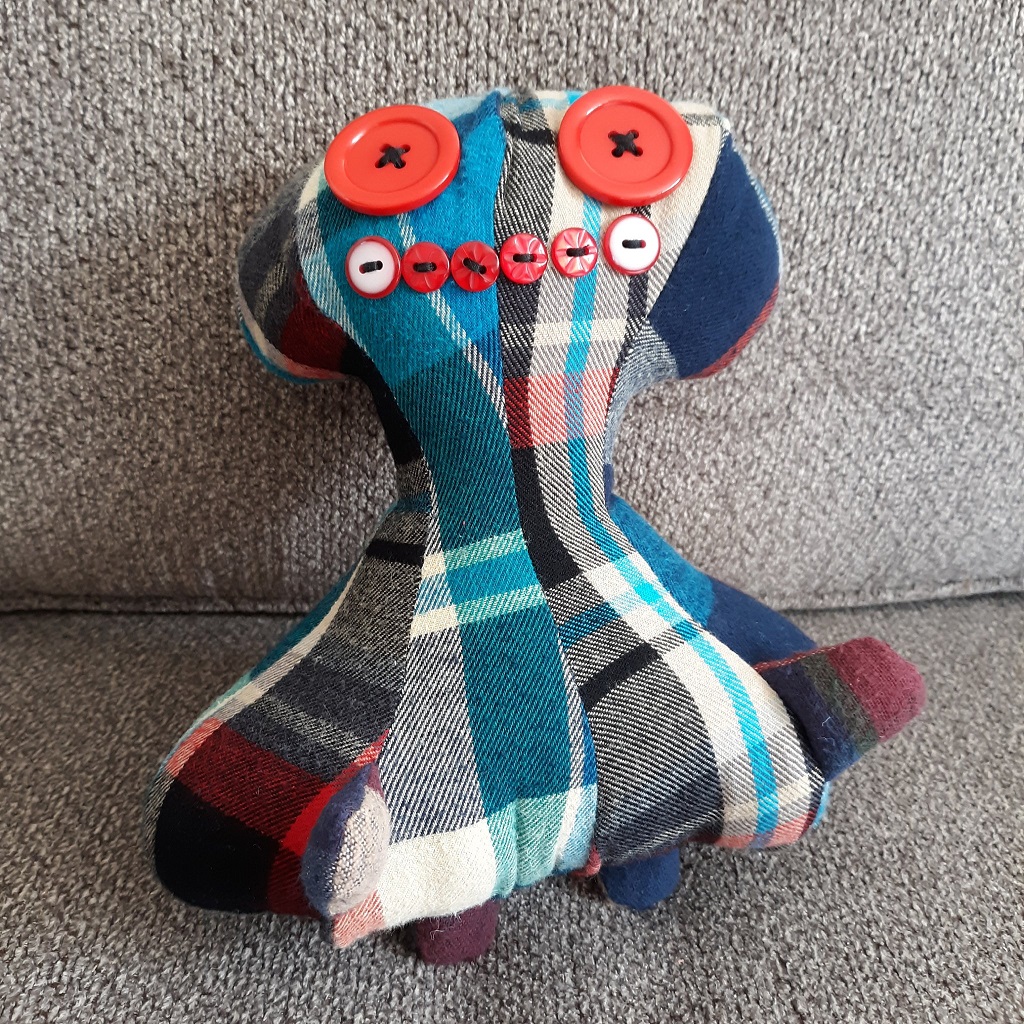 An image of a plaid doll. The doll has a bulbous head with a thin neck and a large, wide body. They kind of look like an octopus. The eyes are made of two red large buttons. The smile is also made of red buttons. The doll has two stubby arms and two stubby legs.