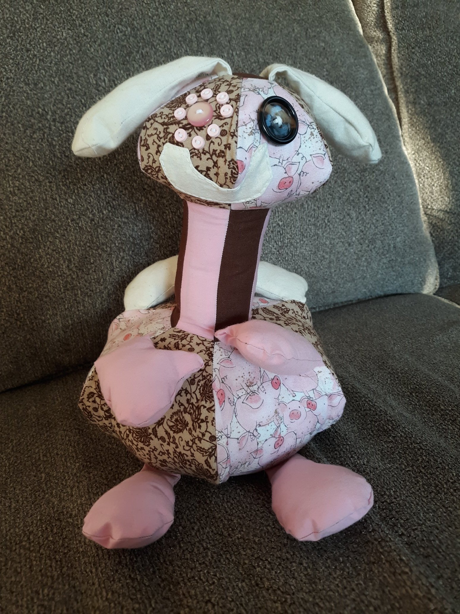 A hand made doll sitting on a couch. It has a round head with alternating floral and pig patterns with a white smile and pink and brown buttons for eyes. There are white ears coming out of the top of the head. The neck is long and thin made of alternating brown and pink fabrics. The body is also round with the same alternating pig and floral patterns. There are pink arms and legs poking out of the body. There are also white wings in the back.