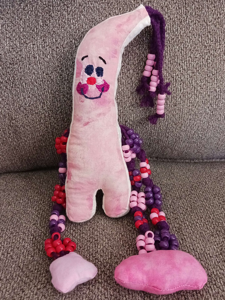 A little pink doll on a couch. They're almost banana shaped but the bottom goes straight down with two stubby legs. They've braided yarn with beads at the ends hanging off the curved tip of their head. They've an embroidered face with two large eyes, a smile, and blushing cheeks. There are two beaded arms with fabric hands on each side of their body.