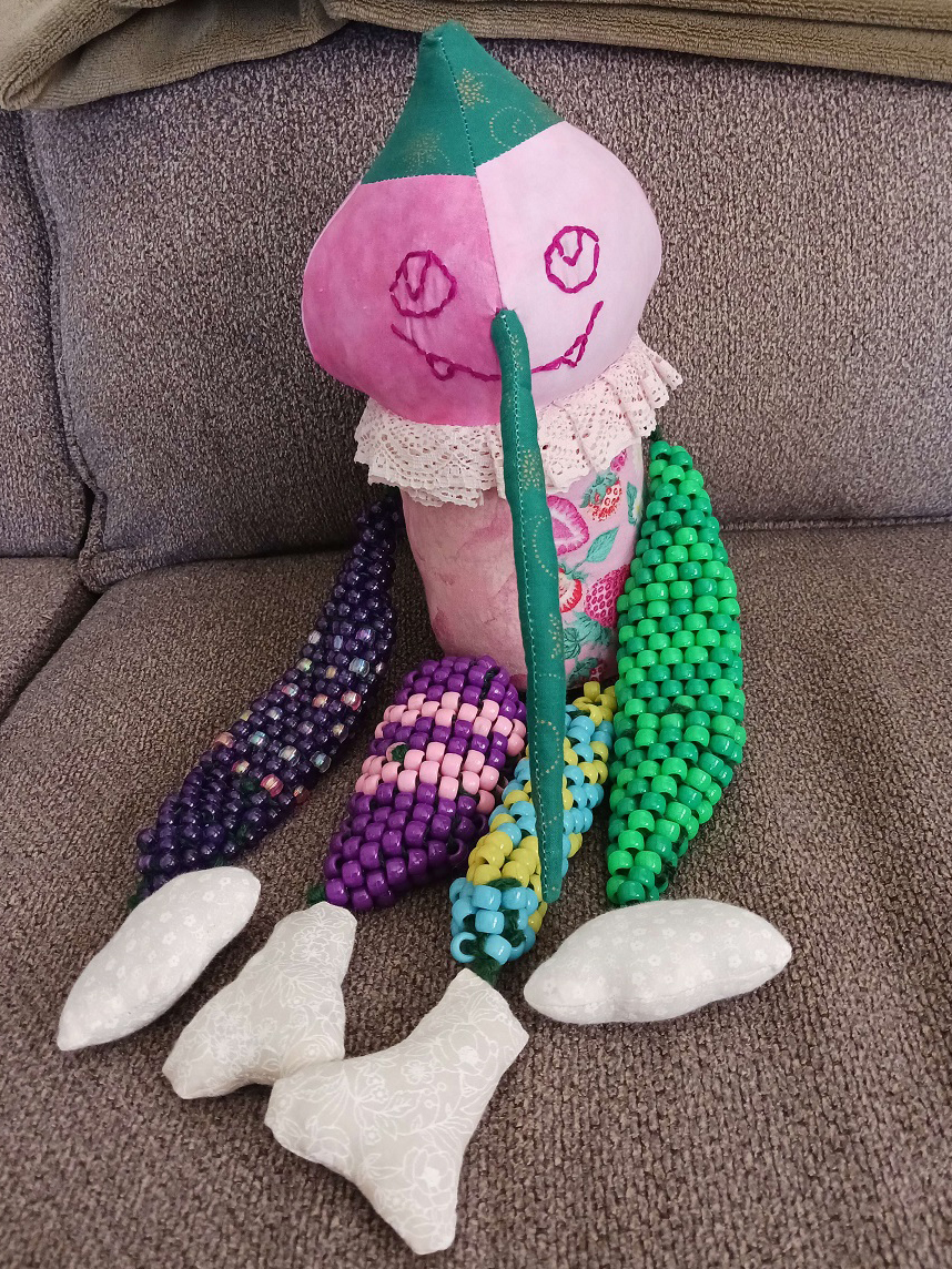 A doll sitting on a couch. They have a pink bulbous head with a green pointed tip on top. Their face is embroidered on with two eyes and a large smile with two pointy teeth. There's a long thin nose protruding from the middle of their face. Their body is tubular with a pink frill at the neck. They have beaded limbs with white fabric hands and feet at the ends.