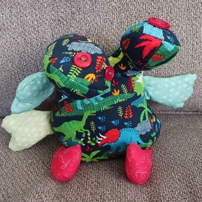 A doll sitting on a couch. It has two heads with buttons for eyes. The heads are attached to a round body with two feet and three arms.