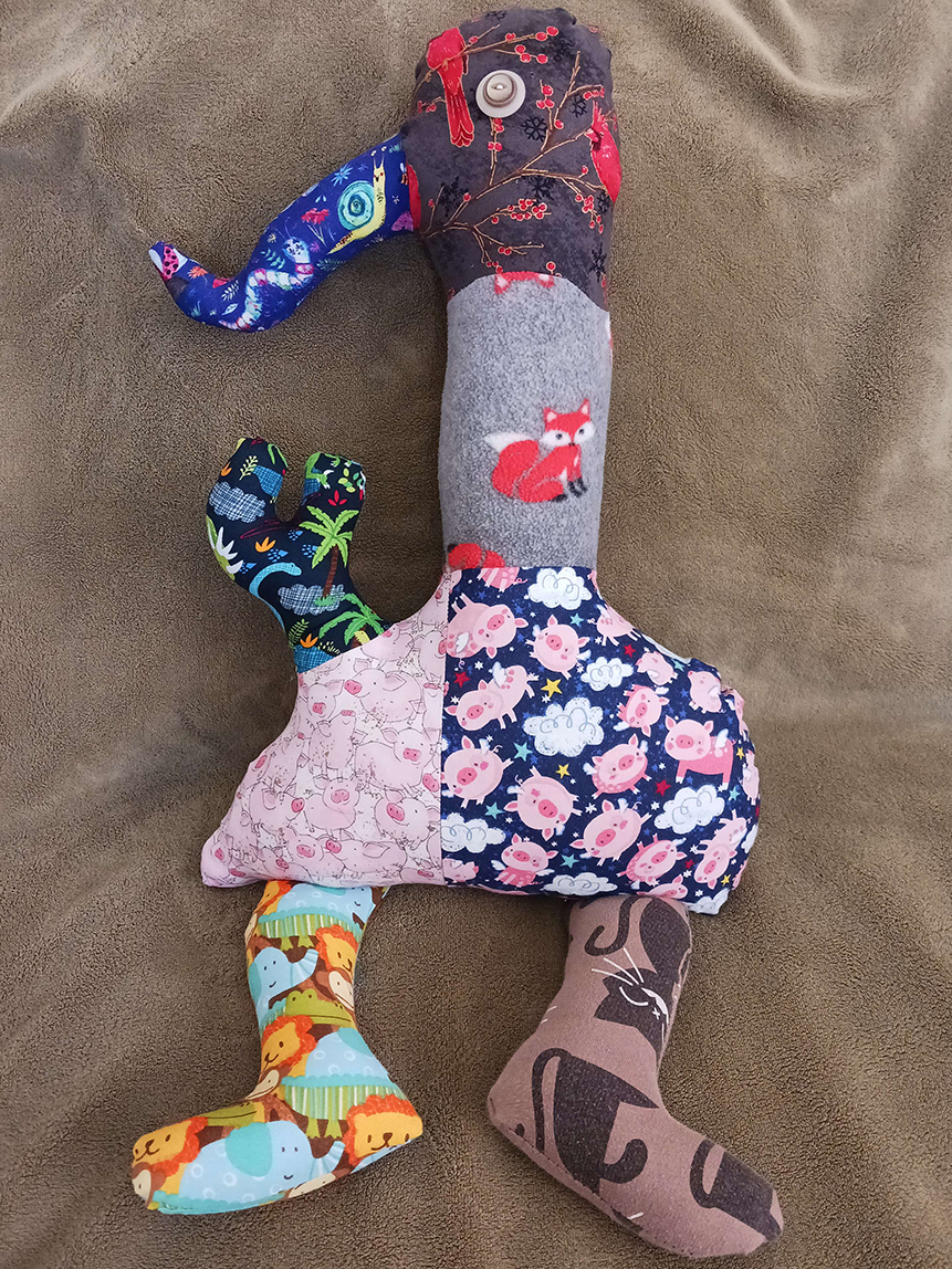 A doll sitting on a couch. It has a long snout coming out of its head with a long neck attached to a round body. On the left is one short arm with two fingers sticking out of the body. It has two legs sticking out from the bottom of the body.