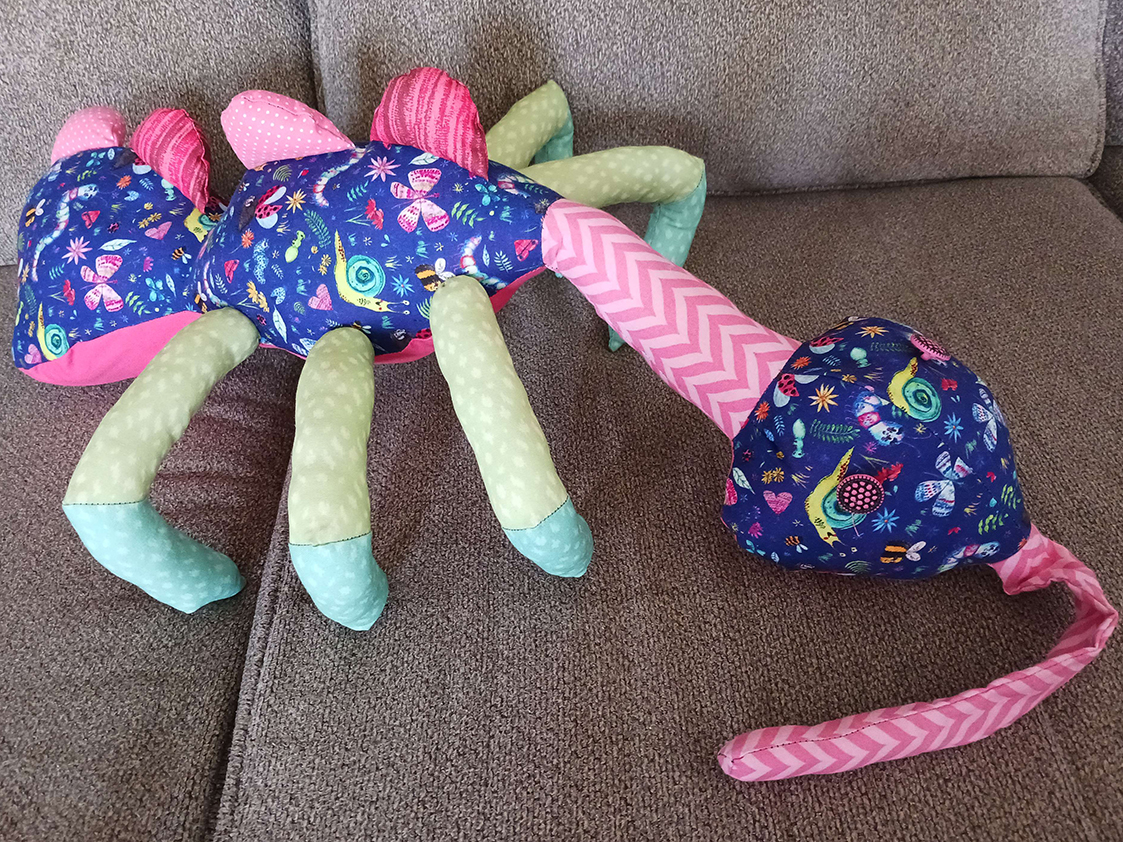 A doll laying on a couch. It looks quite similar to an insect with a head, thorax with three legs on each side, and butt. The head has two button eyes and a long proboscis all on a long neck. The thorax and butt each have two spikes on the top of them.