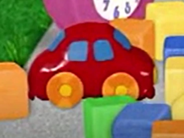A red toy car laying on concrete. There are colorful blocks around it and Tickety is behind it.