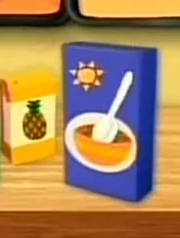 A blue box of cereal sitting on top of a wooden counter. There's an image of a yellow cereal bowl with a white spoon and a sun shining down upon it on the front of the cereal box.
