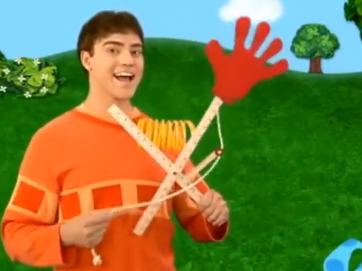 Joe is holding some sort of contraption that has two rulers crossing over each other, a yellow spiral between it, and an orange hand made of paper on the end of one of the rulers. There's a rope attached to the orange hand.