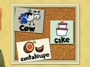 A cork board with a white wooden frame. There are three pieces of paper pinned onto it. One paper has a cow drawn on it, another has a cake, and the third one has a cantaloupe. The cow drawing has a blue paw print on it.