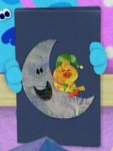 Blue is holding a book that has a little yellow bird laying on top of a crescent moon on the cover of it. The moon is smiling.