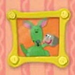 A yellow square frame with swirls in each corner hangs on the wall. The picture inside of it is of Green Kangaroo holding up a rock. The background of the picture is orange.