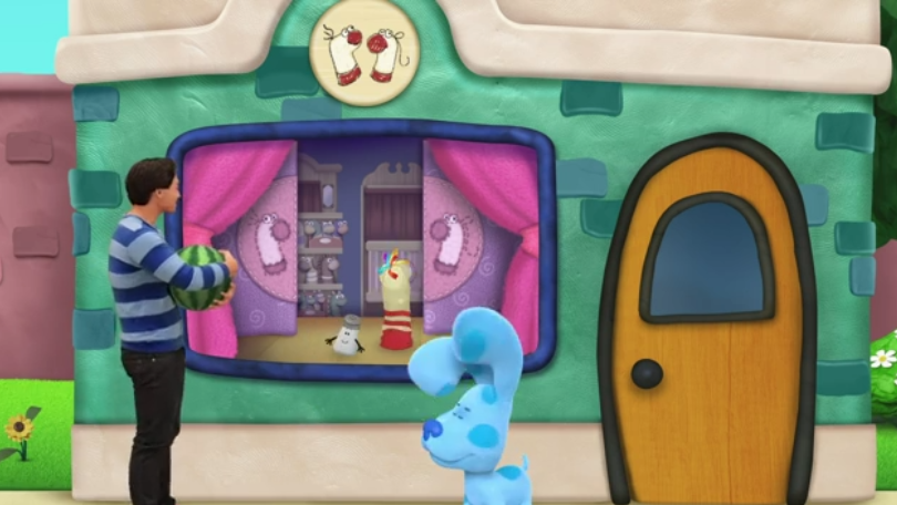 Josh and Blue are standing in front of a green puppet store. Mr. Salt is with a sock puppet behind the window.