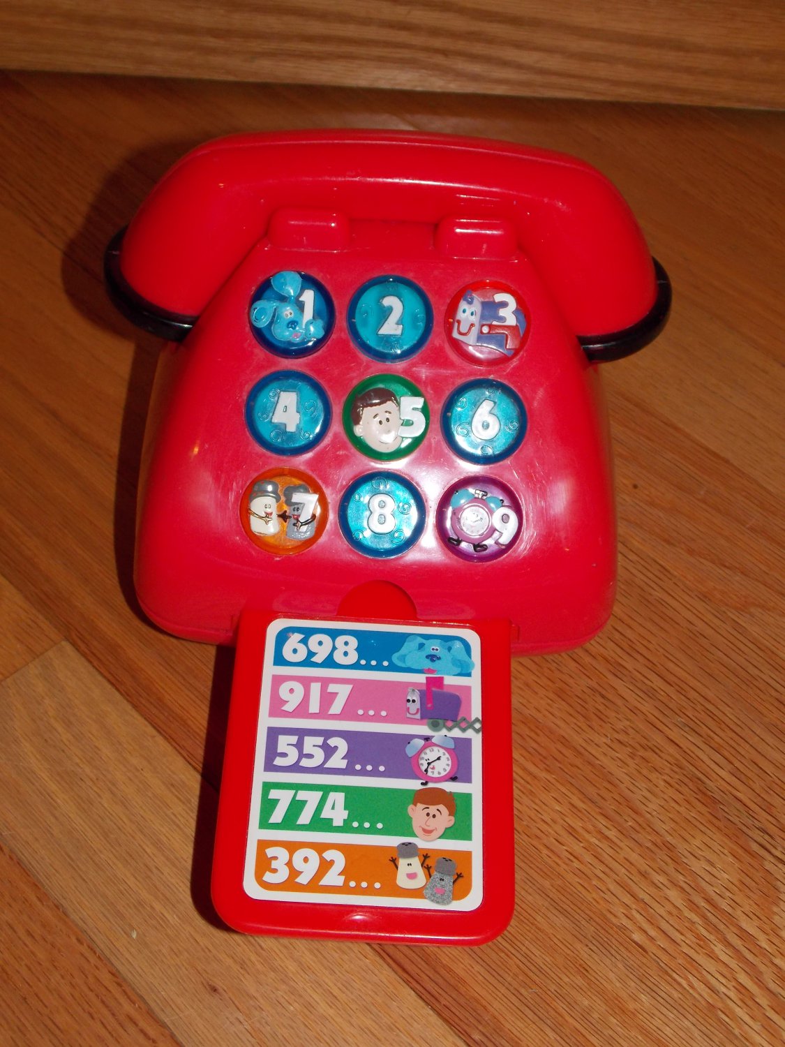 An image of a red toy phone. The odd numbers on the phone have a Blue's Clues character on it. 1 has Blue on it, 3 has Mailbox on it, 5 has Steve on it, 7 has Mr. Salt and Mrs. Pepper on it, and 9 has Tickety Tock on it. A flat tray is pulled out of the bottom of the phone and has an image of different numbers that correspond with different characters. Blue is on the top with the number 698, below her is Mailbox with the number 917, below him is Tickety with the number 552, below her is Steve with the number 774, and below him is Mr. Salt and Mrs. Pepper with the number 392.