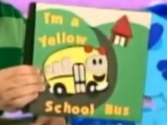 Steve is holding a book that is titled 'I'm a Yellow School Bus.' The cover has an image of a yellow school bus on a road that is between two green hills and has a red building in the background.