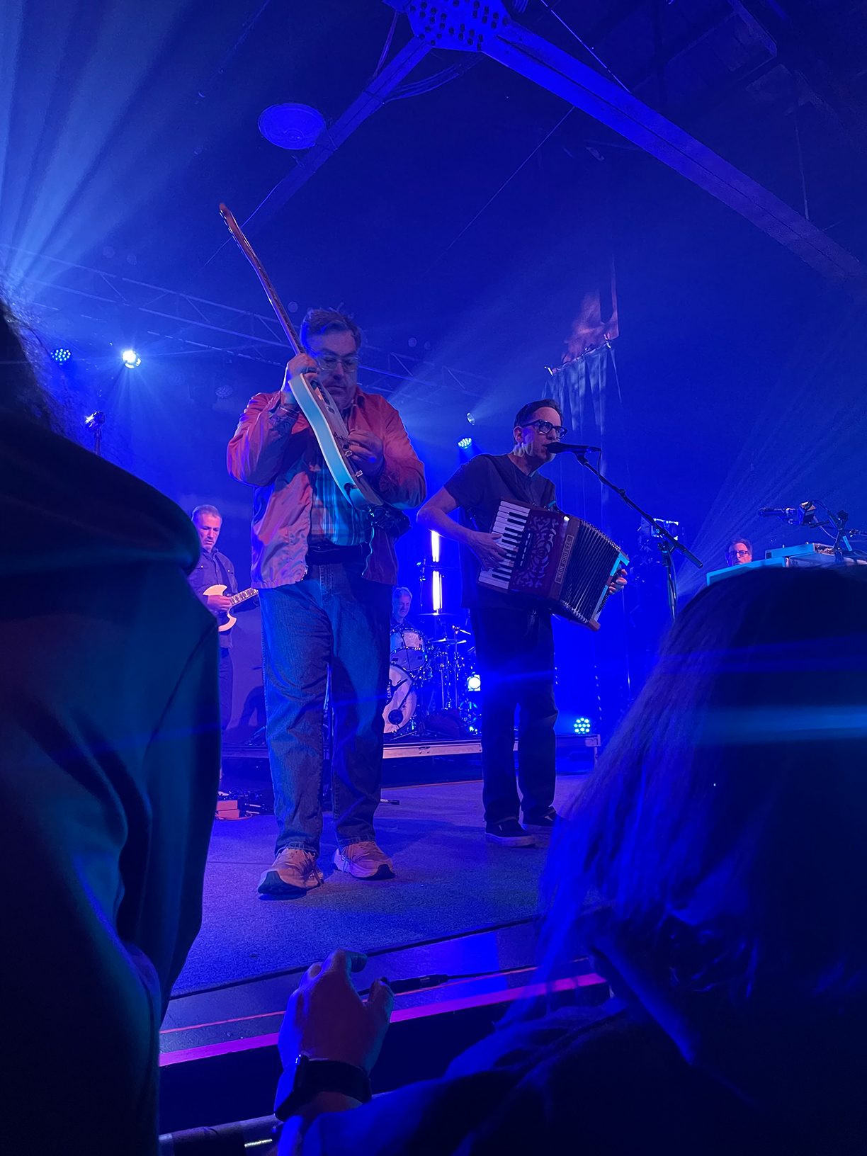 John Flansburgh and John Linnell on stage. Flansburgh is playing his guitar whilst Linnell is playing an accordion and singing into a microphone.