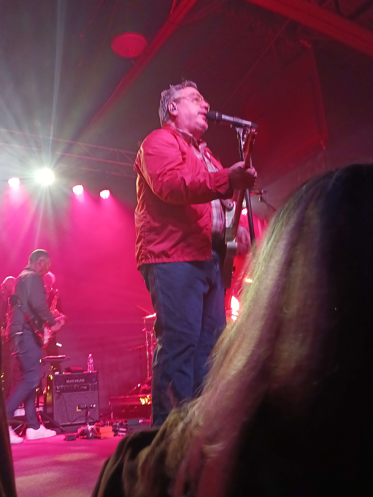 John Flansburgh on stage playing a guitar whilst singing into a microphone.