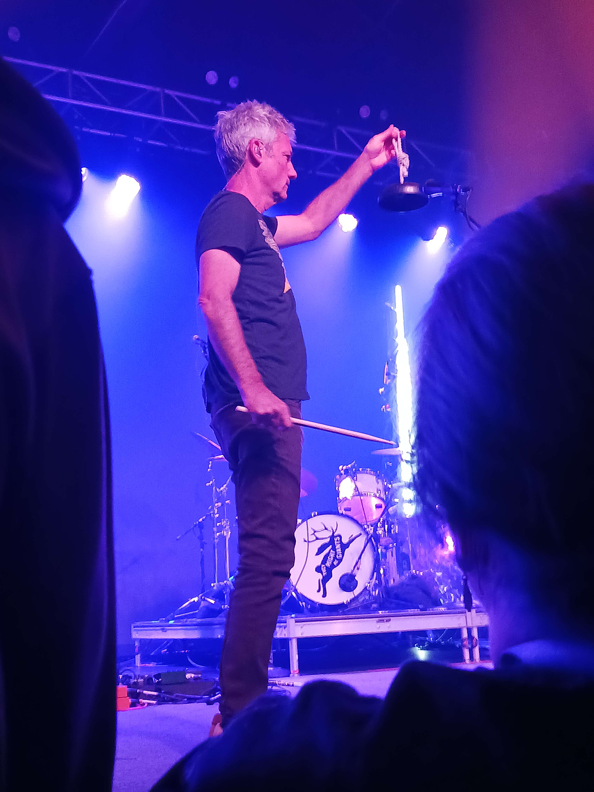 Marty Beller at the front of the stage holding up a bell with a drumstick in the other hand.