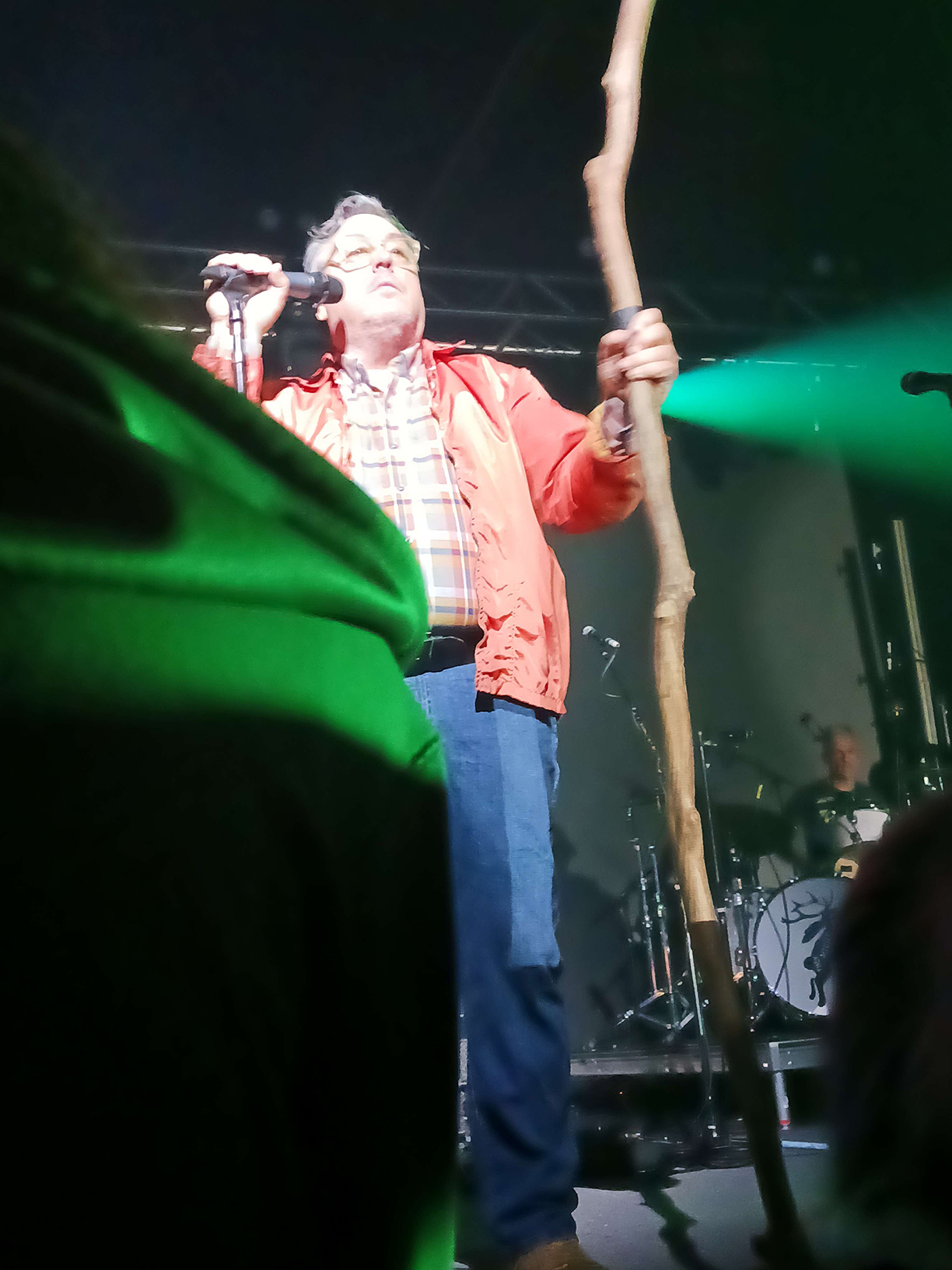 John Flansburgh on stage at his microphone holding a big stick.