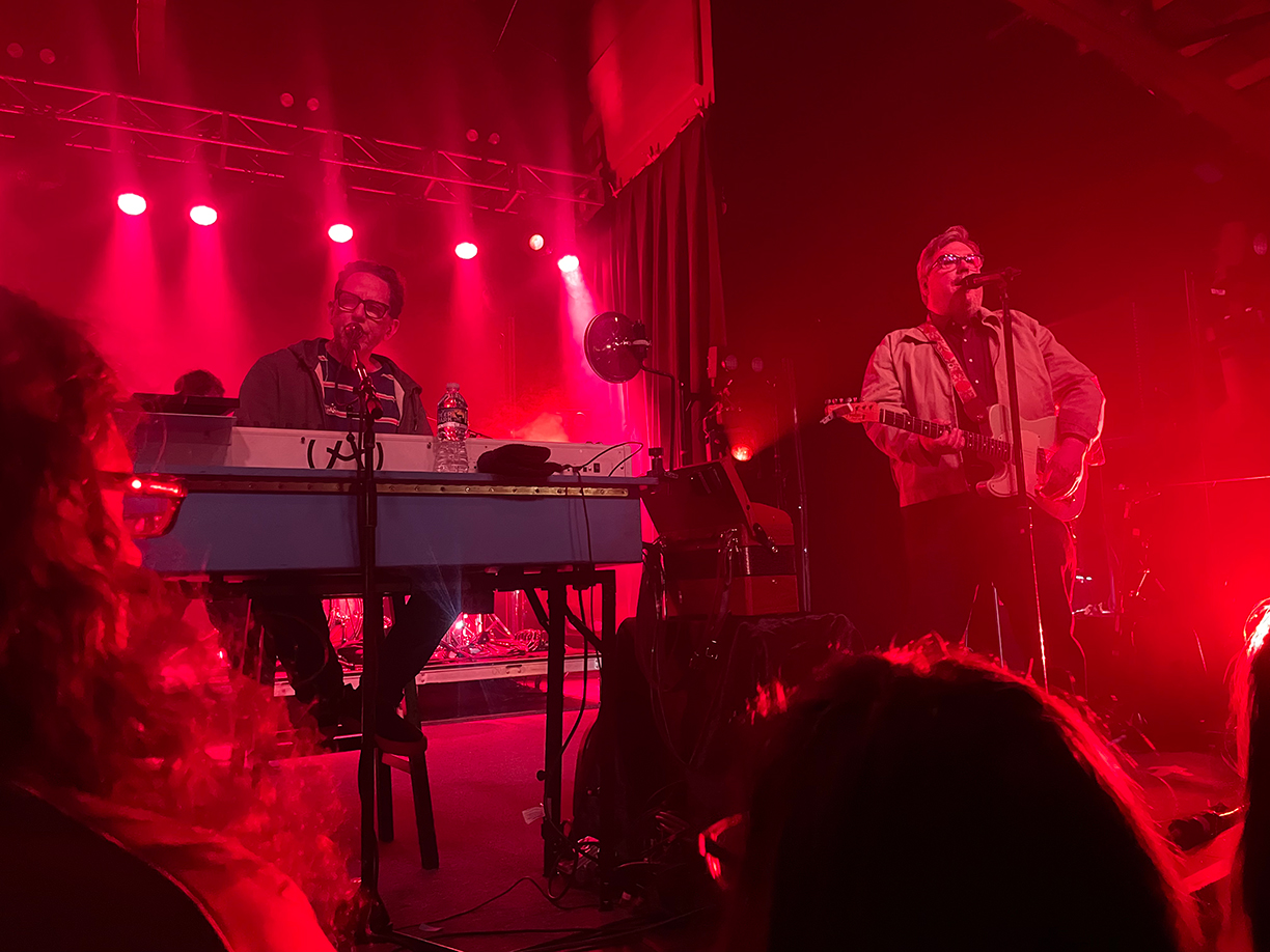 John Linnell and John Flansburgh on stage. Linnell is at his keyboard singing into a microphone. Flansburgh is playing a guitar whilst singing into a microphone.