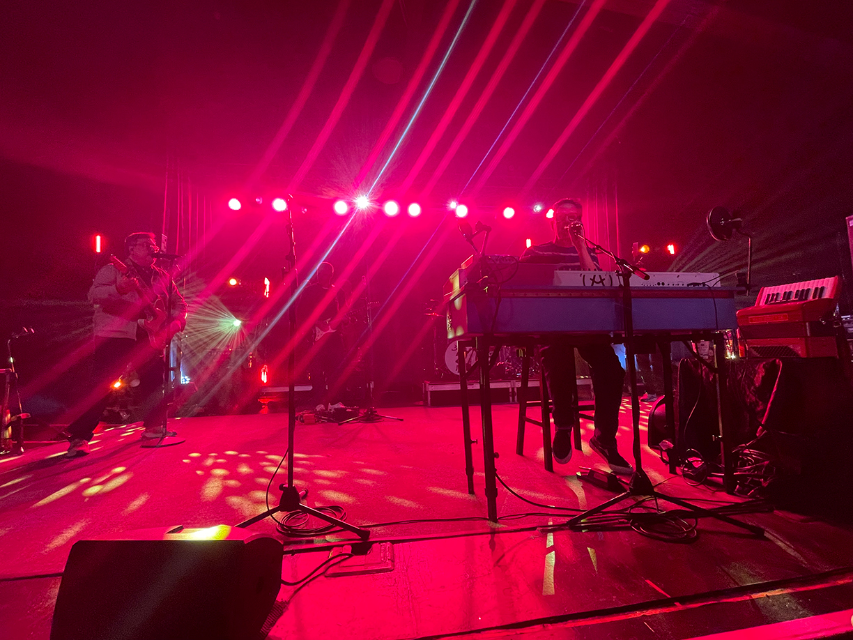 John Linnell and John Flansburgh on stage. Flansburgh is playing a guitar whilst singing into a microphone and Linnell is at his keyboard also singing into a microphone.> <img src=