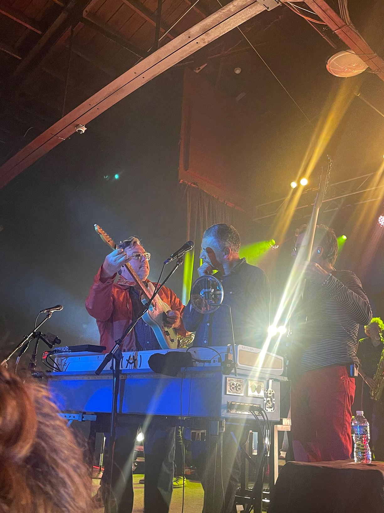 John Flansburgh, Dan Miller, and Danny Weinkauf on stage all standing behind the keyboard. Flansburgh is playing guitar and Weinkauf is playing bass guitar with them standing on each side of Miller.