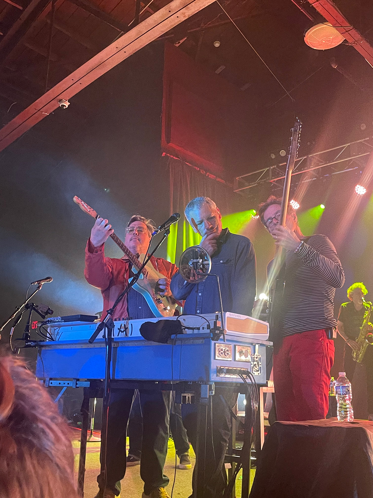 John Flansburgh, Dan Miller, and Danny Weinkauf on stage all standing behind the keyboard. Flansburgh is playing guitar and Weinkauf is playing bass guitar with them standing on each side of Miller.