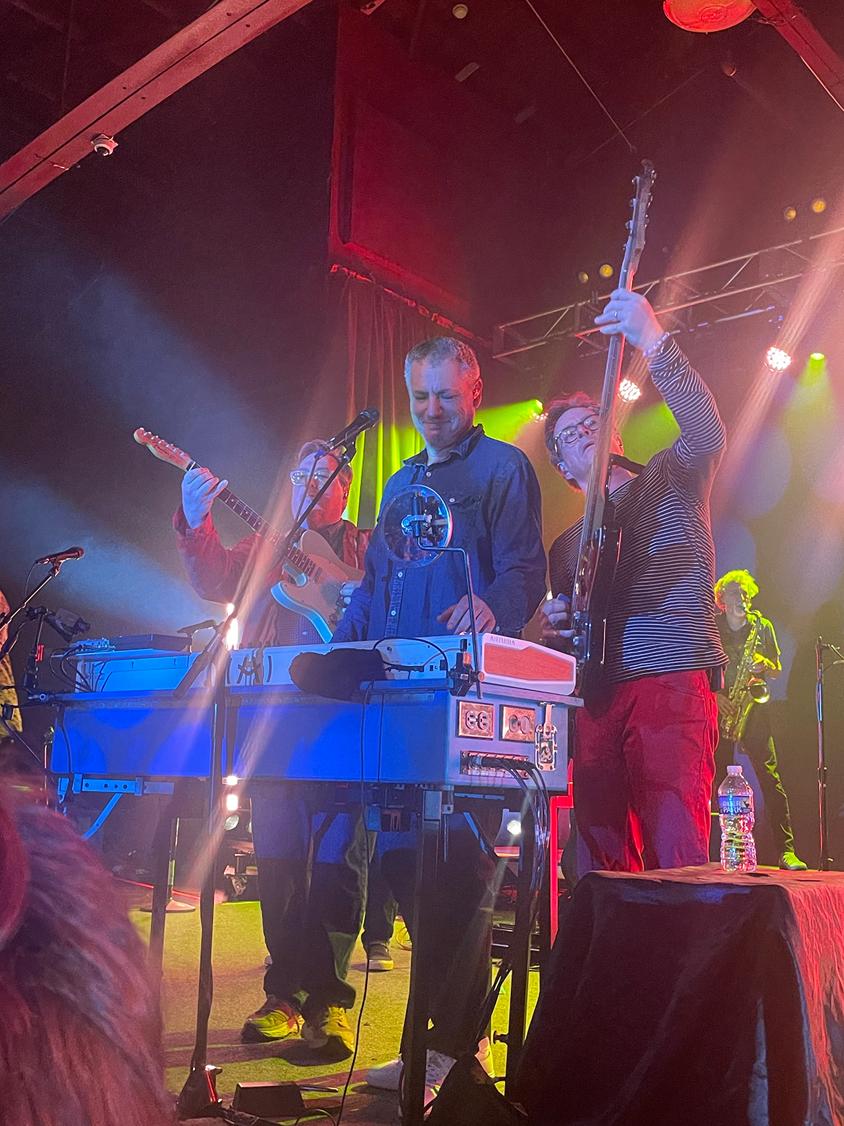 John Flansburgh, Dan Miller, and Danny Weinkauf on stage all standing behind the keyboard. Flansburgh is playing guitar and Weinkauf is playing bass guitar with them standing on each side of Miller.