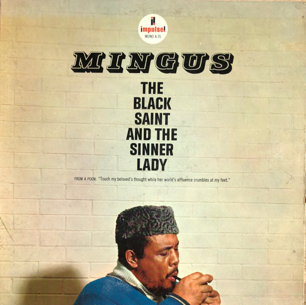 Album cover of The Black Saint and the Sinner Lady by Charles Mingus. Mingus is down below lighting a pipe in front of a white bricked wall. The album title is above him.