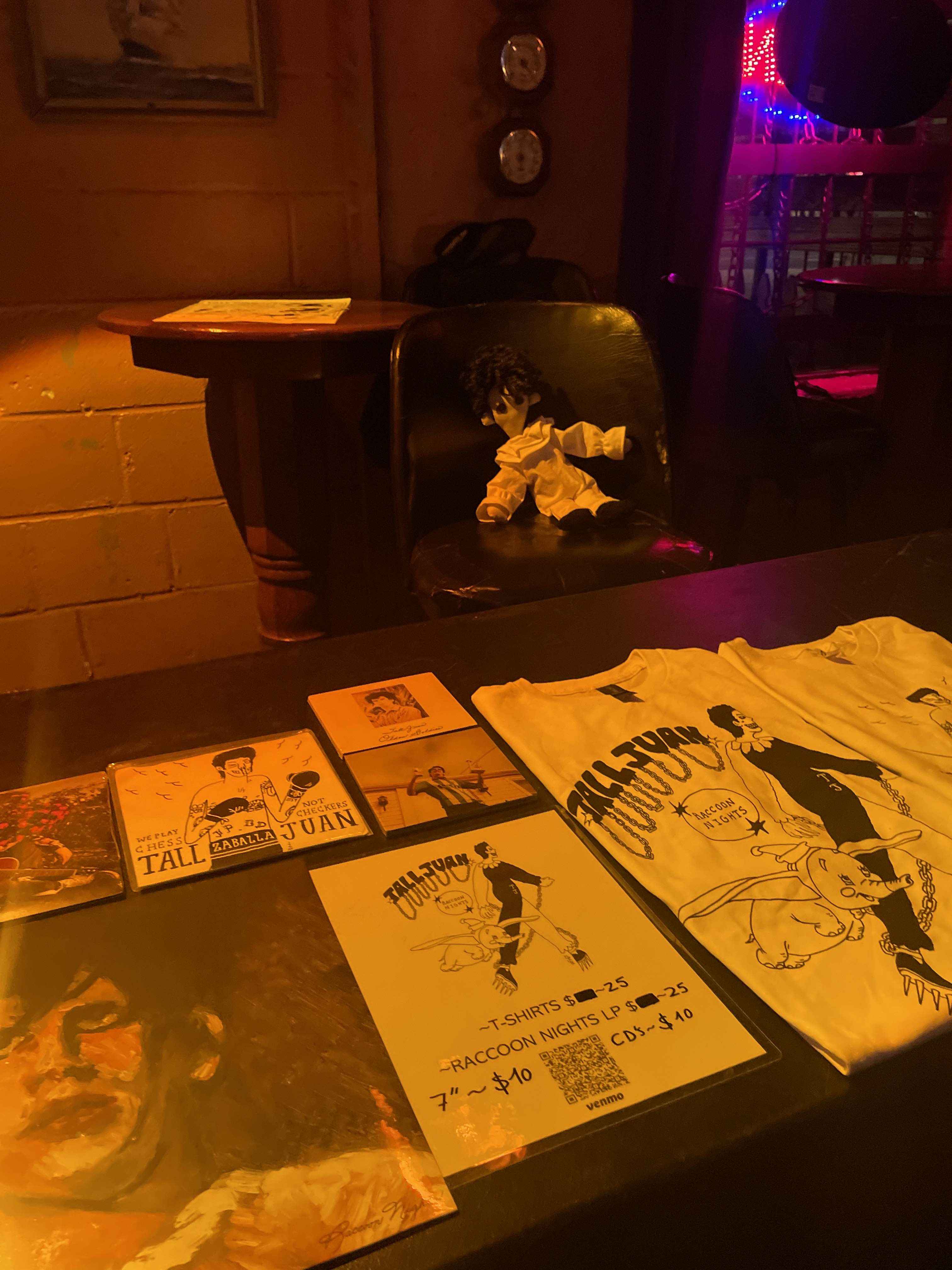 A doll of Tall Juan sitting on a chair behind a table full of Tall Juan merchandise.