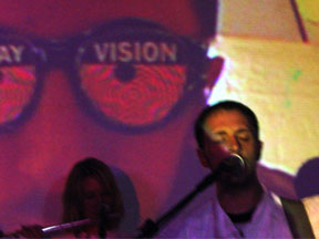 Burns singing into the microphone with an image of him wearing x-ray goggles behind him.