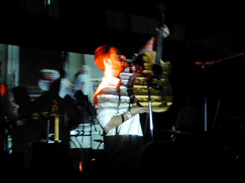 Burns holding up his guitar whilst singing into the microphone. There are images projected over him.