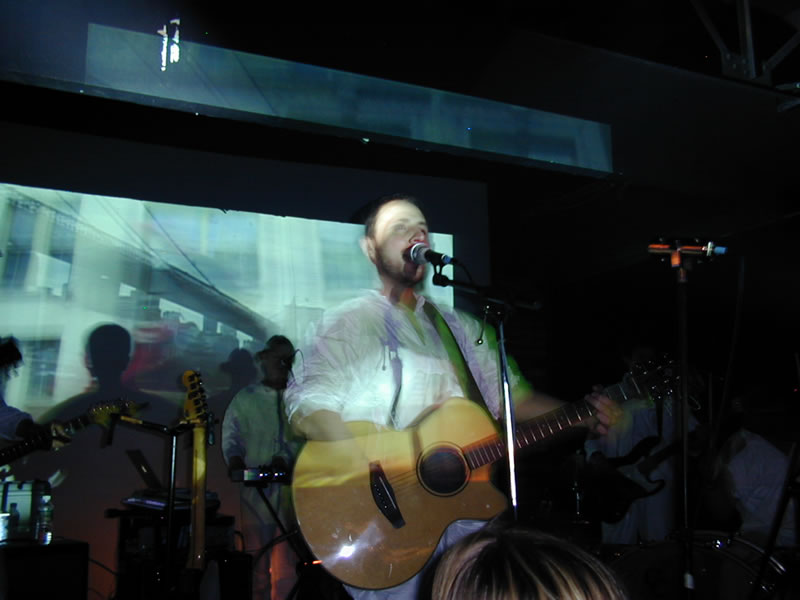 Burns singing whilst playing guitar. There are images projected over him.